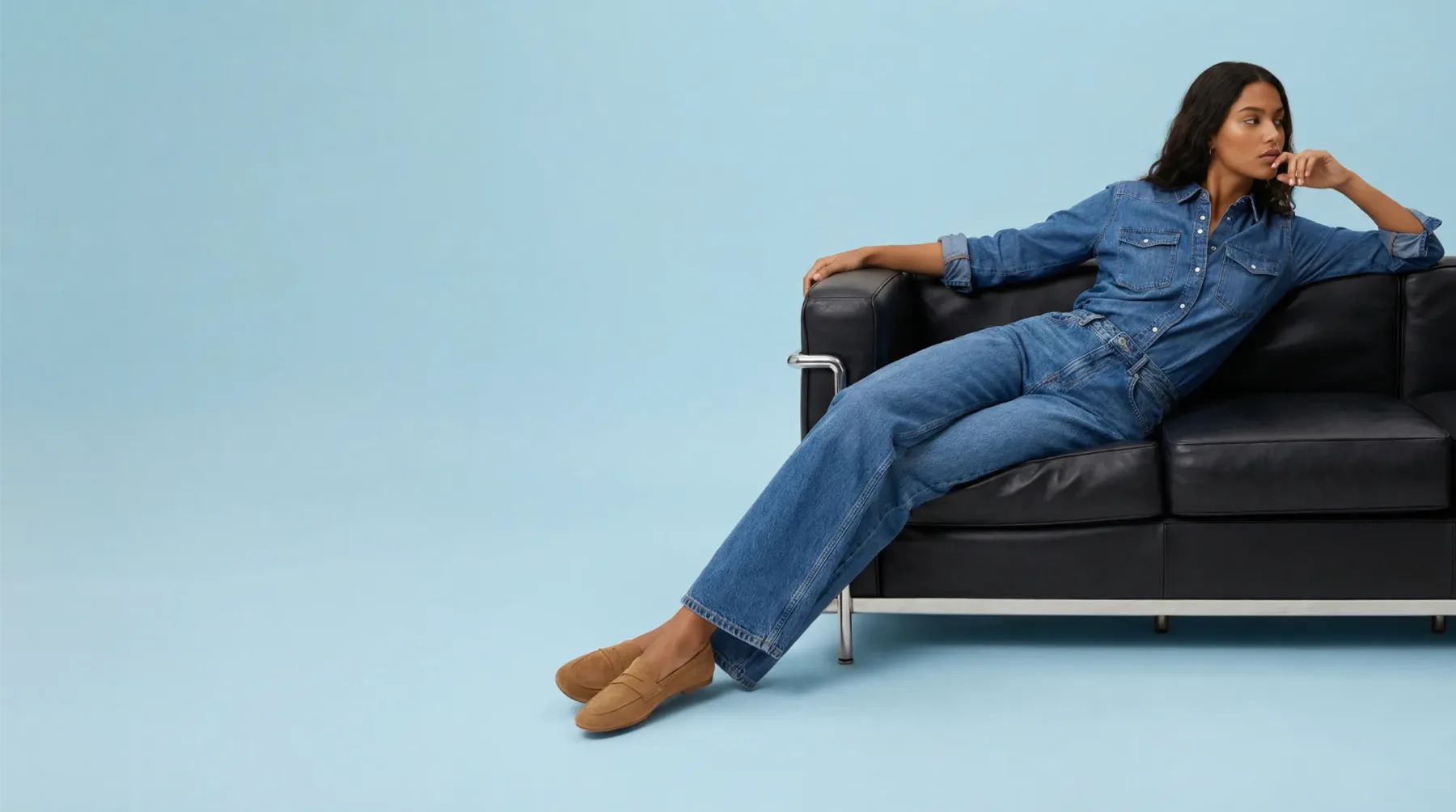 Modella seduta su divano nero con outfit denim completo su sfondo azzurro pastello, immagine fashion e-commerce generata con AI