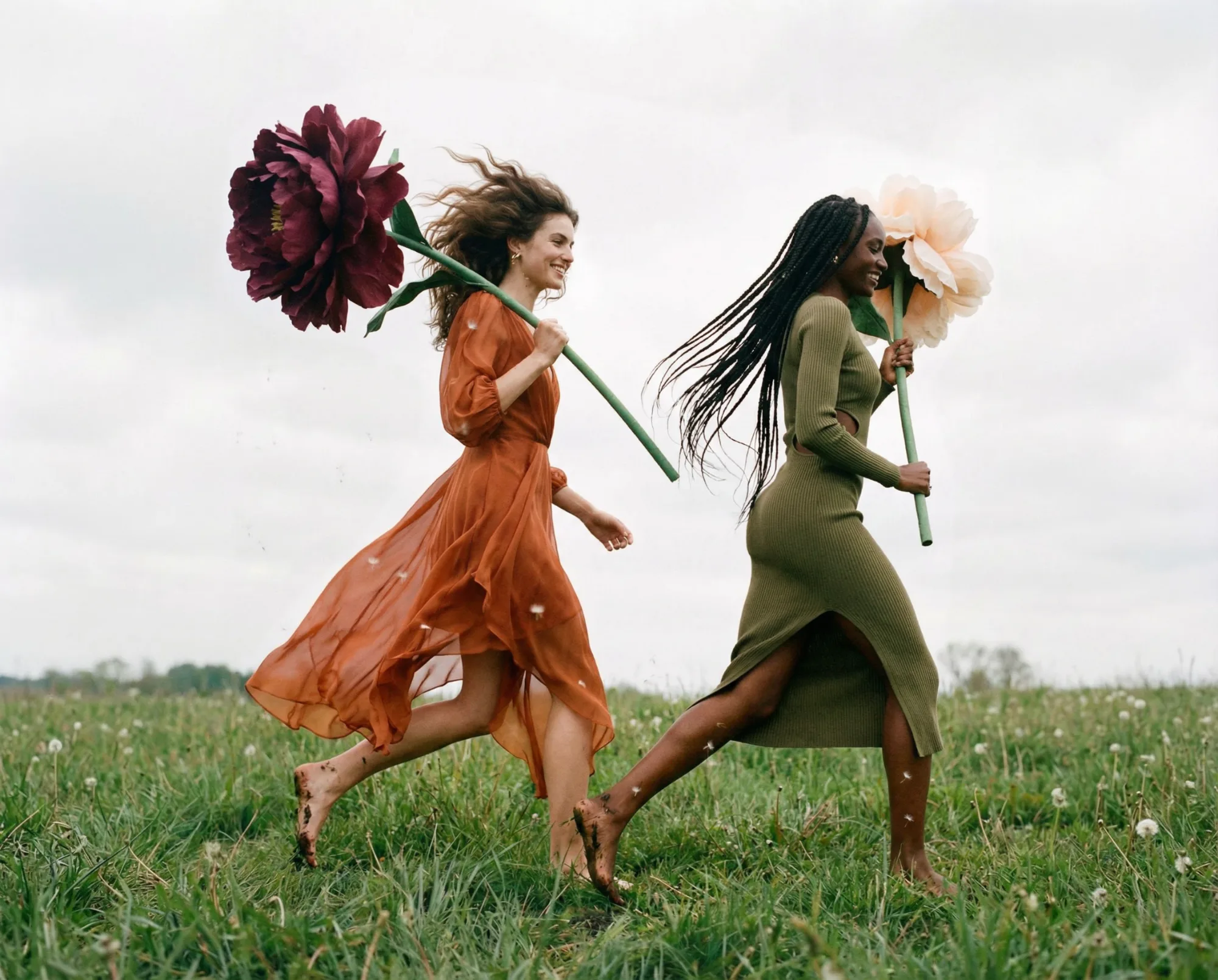 Due donne che corrono in un prato indossando abiti colorati mentre tengono grandi fiori.
