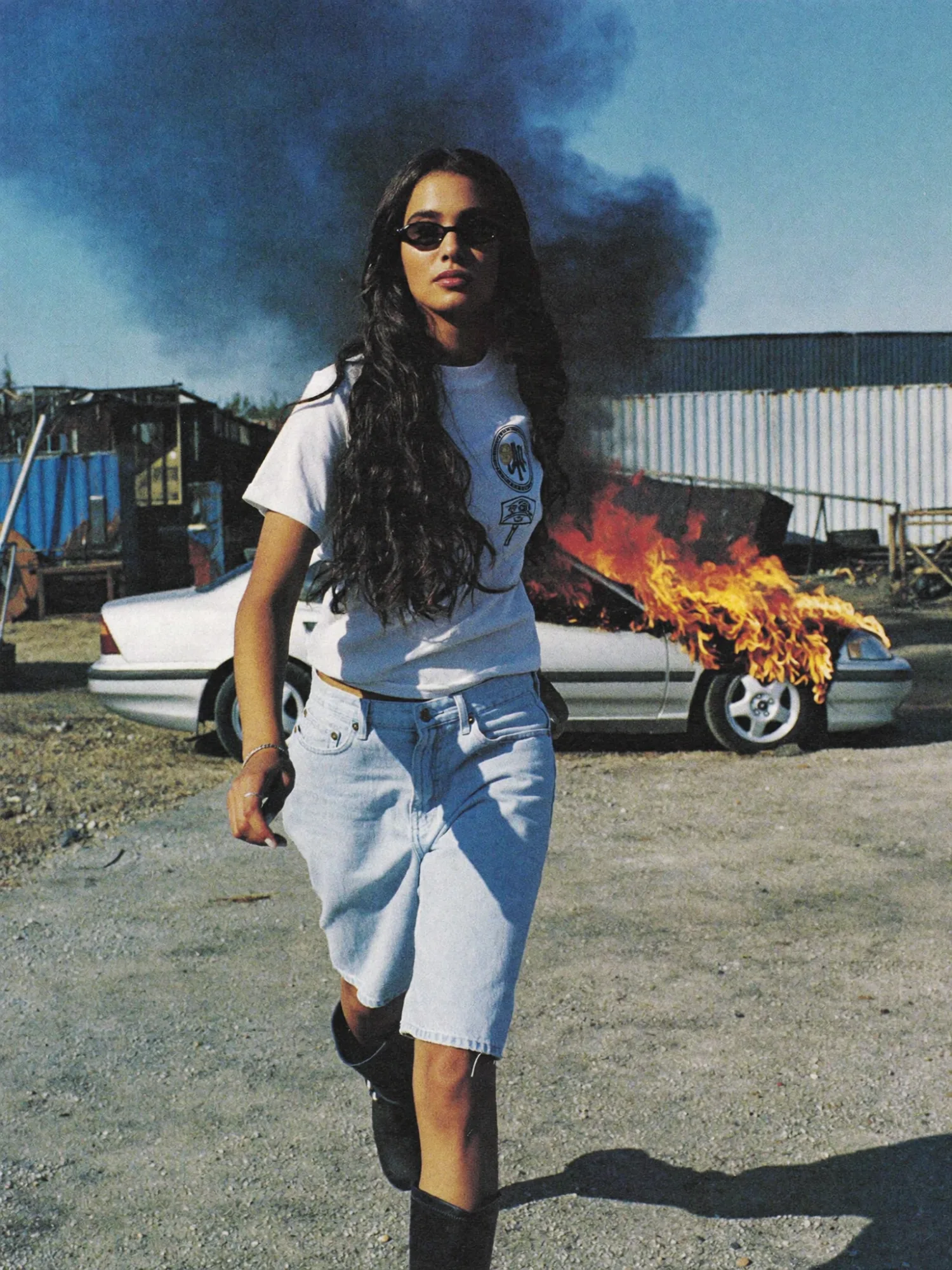 Modella con occhiali da sole e outfit casual che cammina in un ambiente industriale mentre un’auto in fiamme brucia sullo sfondo.