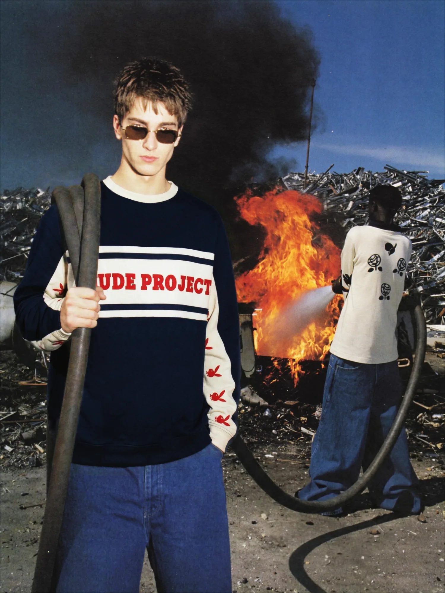 Modello con occhiali da sole e maglia streetwear davanti a un incendio in una discarica industriale, mentre un uomo sullo sfondo usa un tubo per spegnere il fuoco.