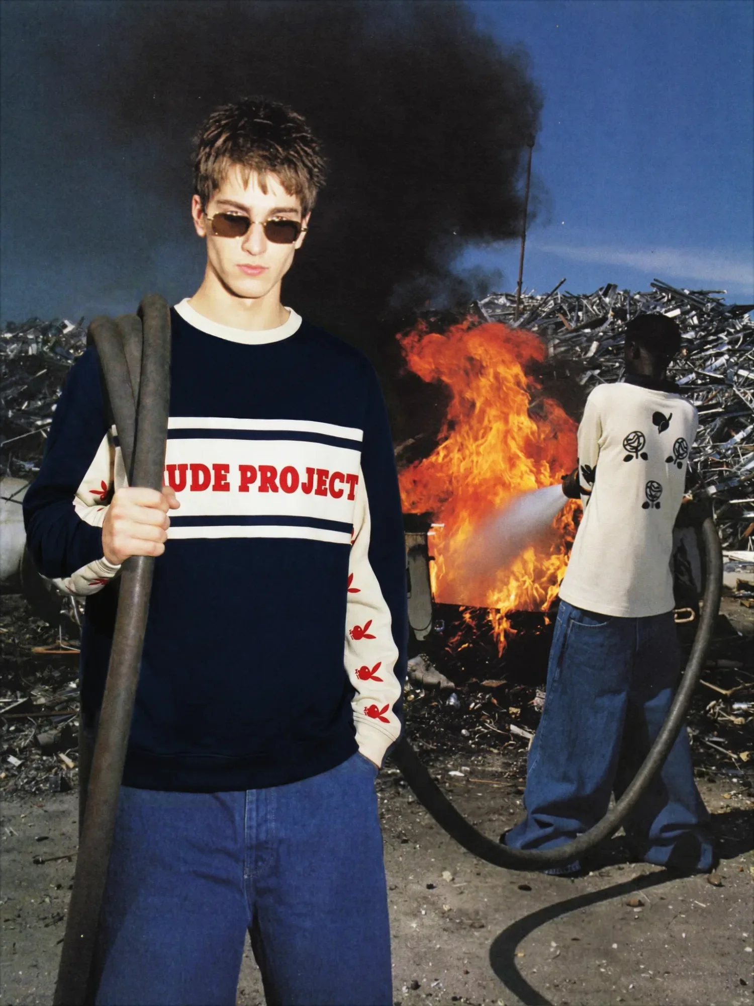 Modello con occhiali da sole e maglia streetwear davanti a un incendio in una discarica industriale, mentre un uomo sullo sfondo usa un tubo per spegnere il fuoco.