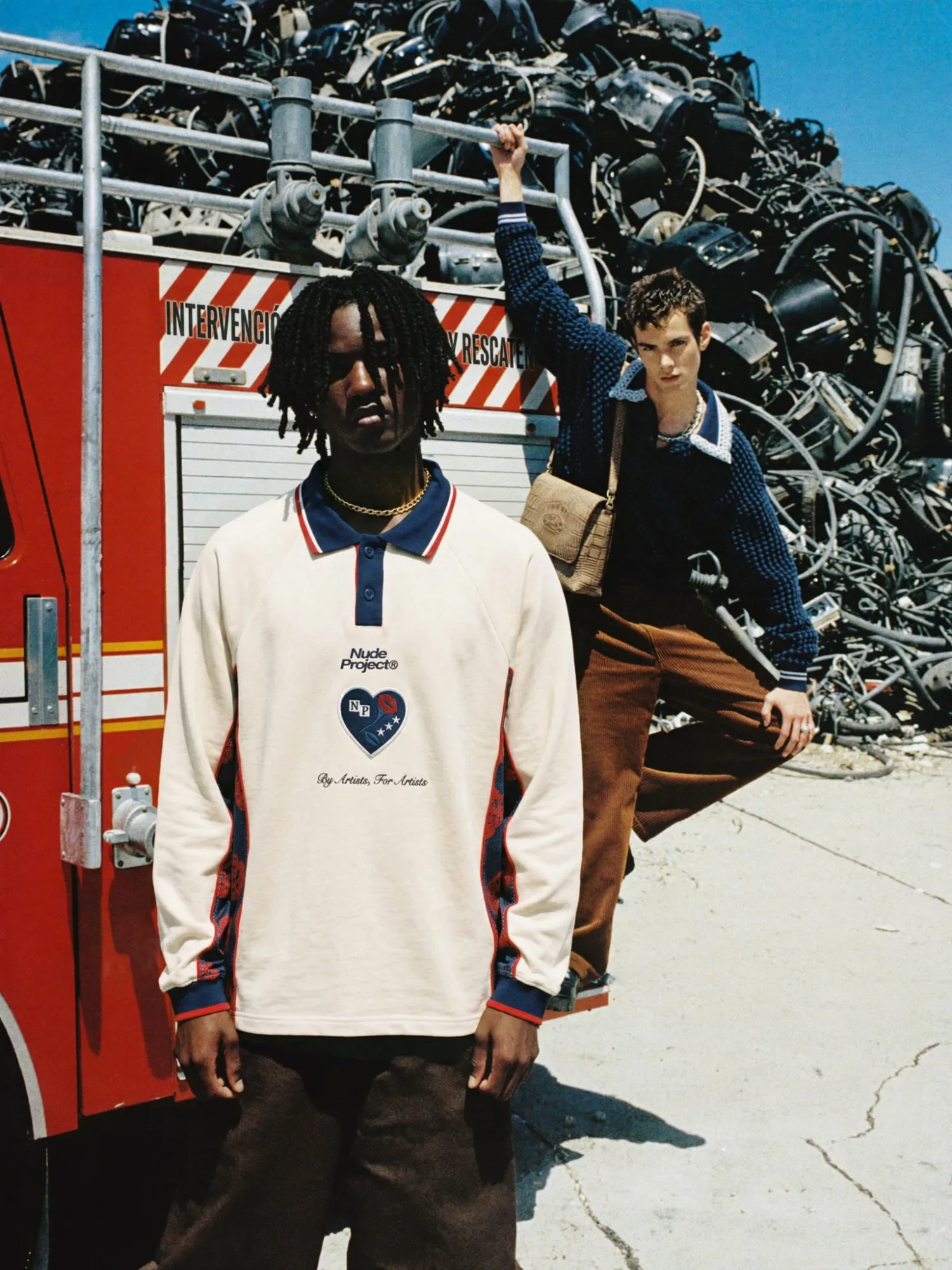 Due modelli in abbigliamento streetwear davanti a un camion dei vigili del fuoco e a una grande pila di rottami metallici in un ambiente industriale.