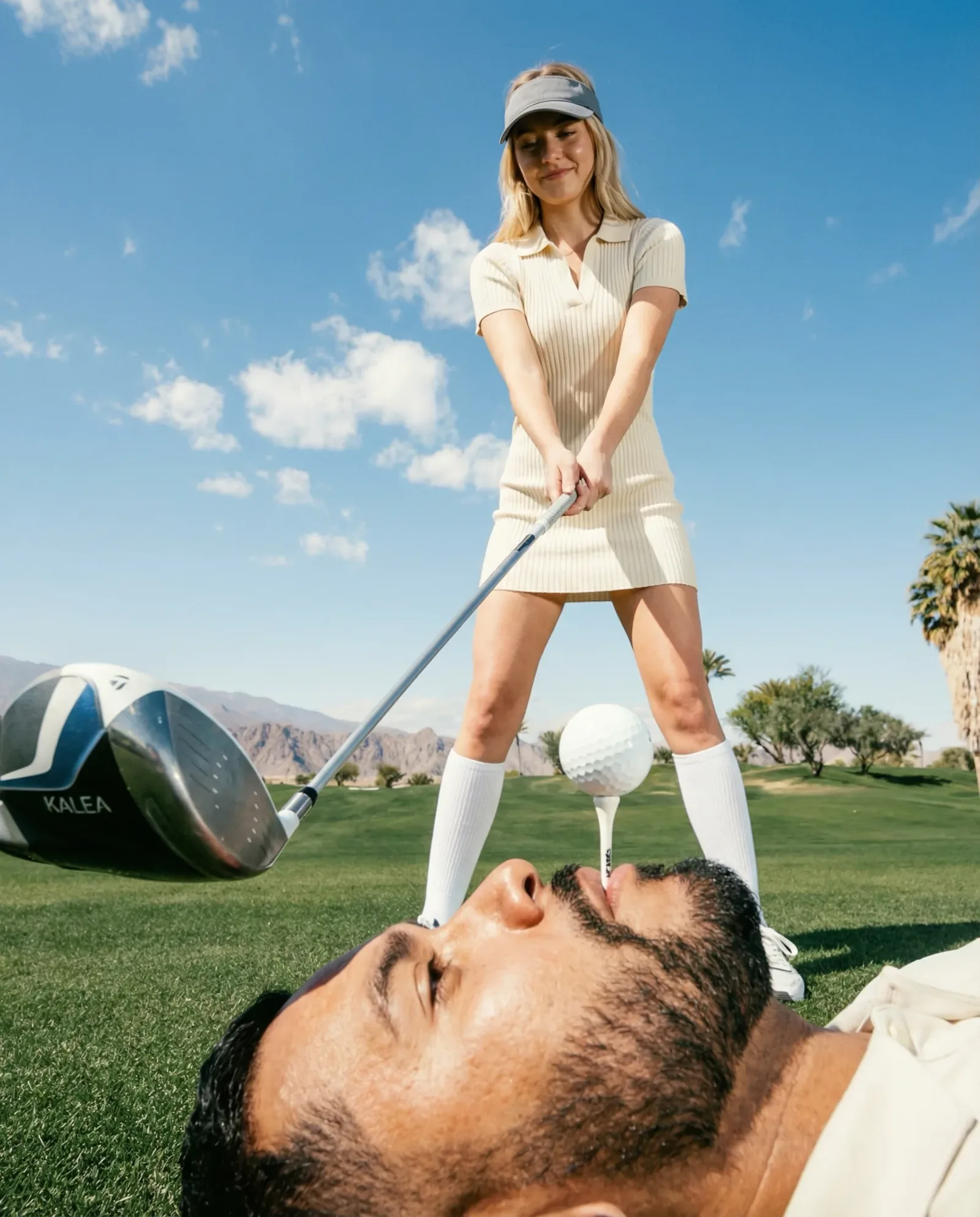 Scena creativa su un campo da golf con prospettiva forzata: una golfista pronta a colpire la pallina posizionata sulla bocca di un uomo sdraiato sull’erba.