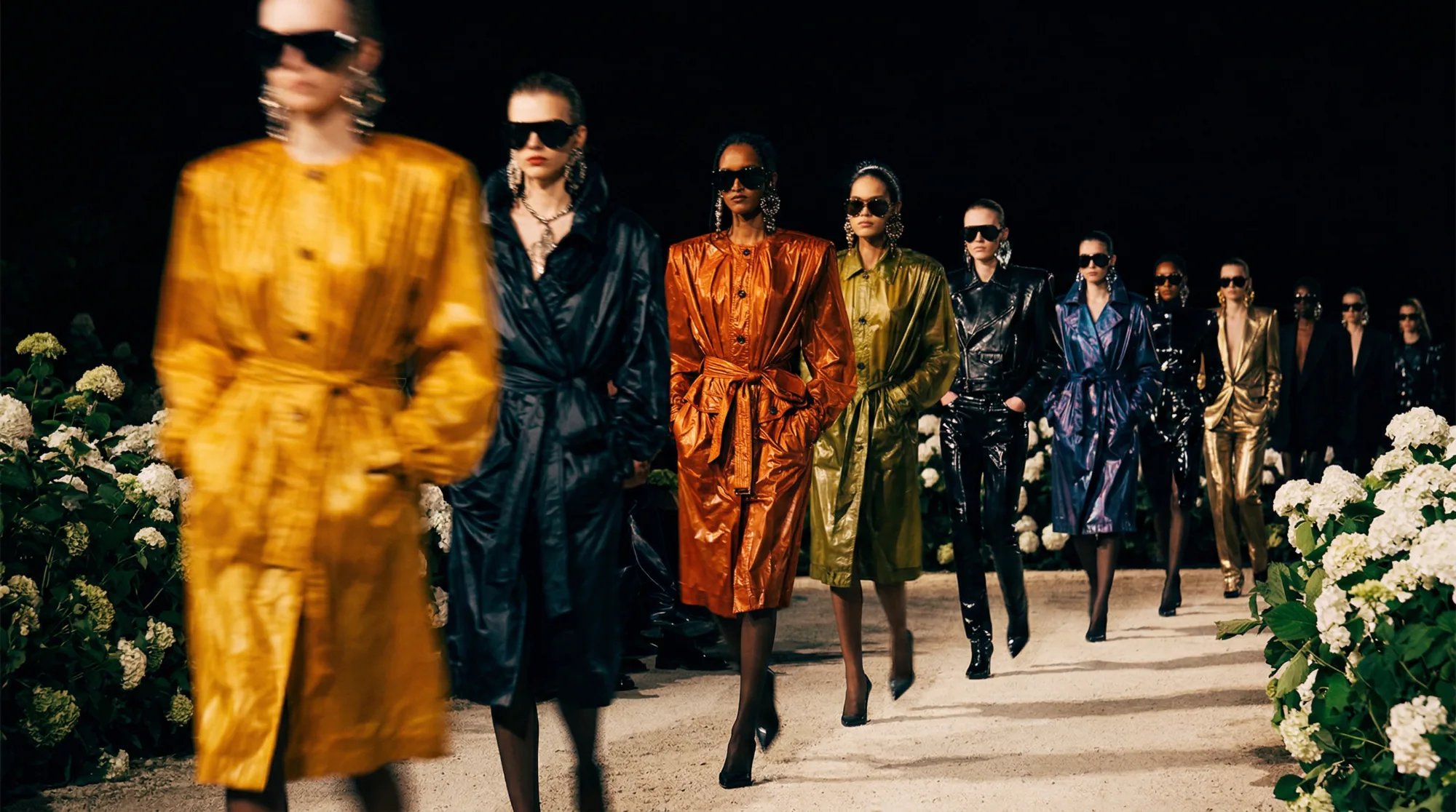 Sfilata di moda notturna con modelle che indossano trench lucidi colorati su passerella circondata da fiori bianchi.