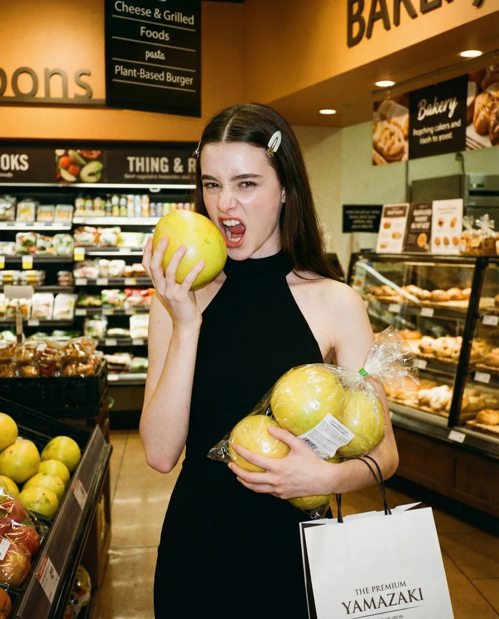 Donna in abito nero nel reparto frutta di un supermercato mentre tiene delle mele e ne morde una.  English (alt):