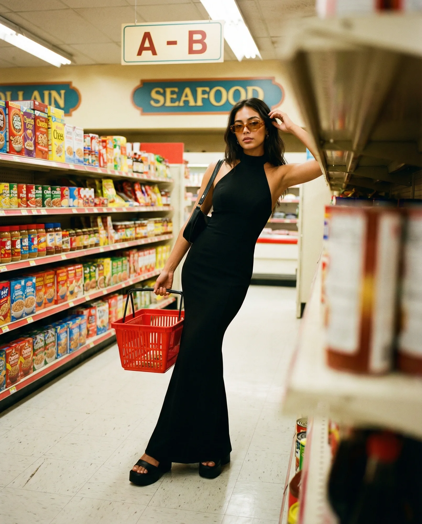 Donna in abito nero che cammina tra gli scaffali di un supermercato con un cestino della spesa rosso.