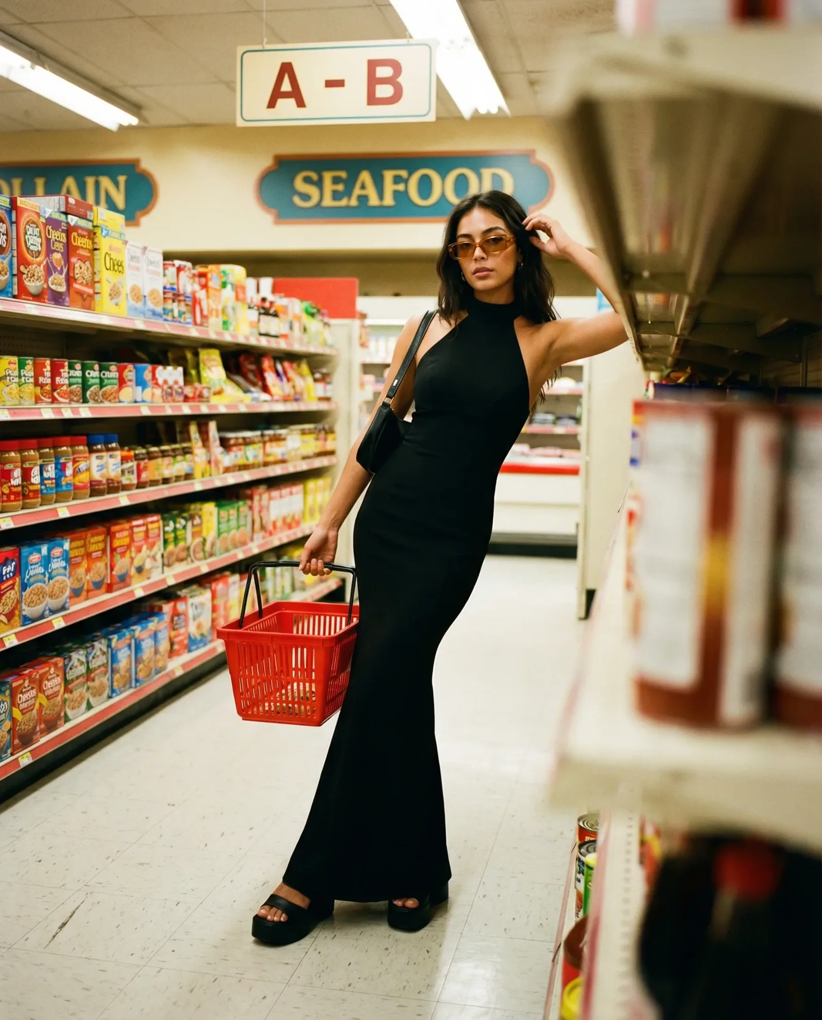 Donna in abito nero che cammina tra gli scaffali di un supermercato con un cestino della spesa rosso.