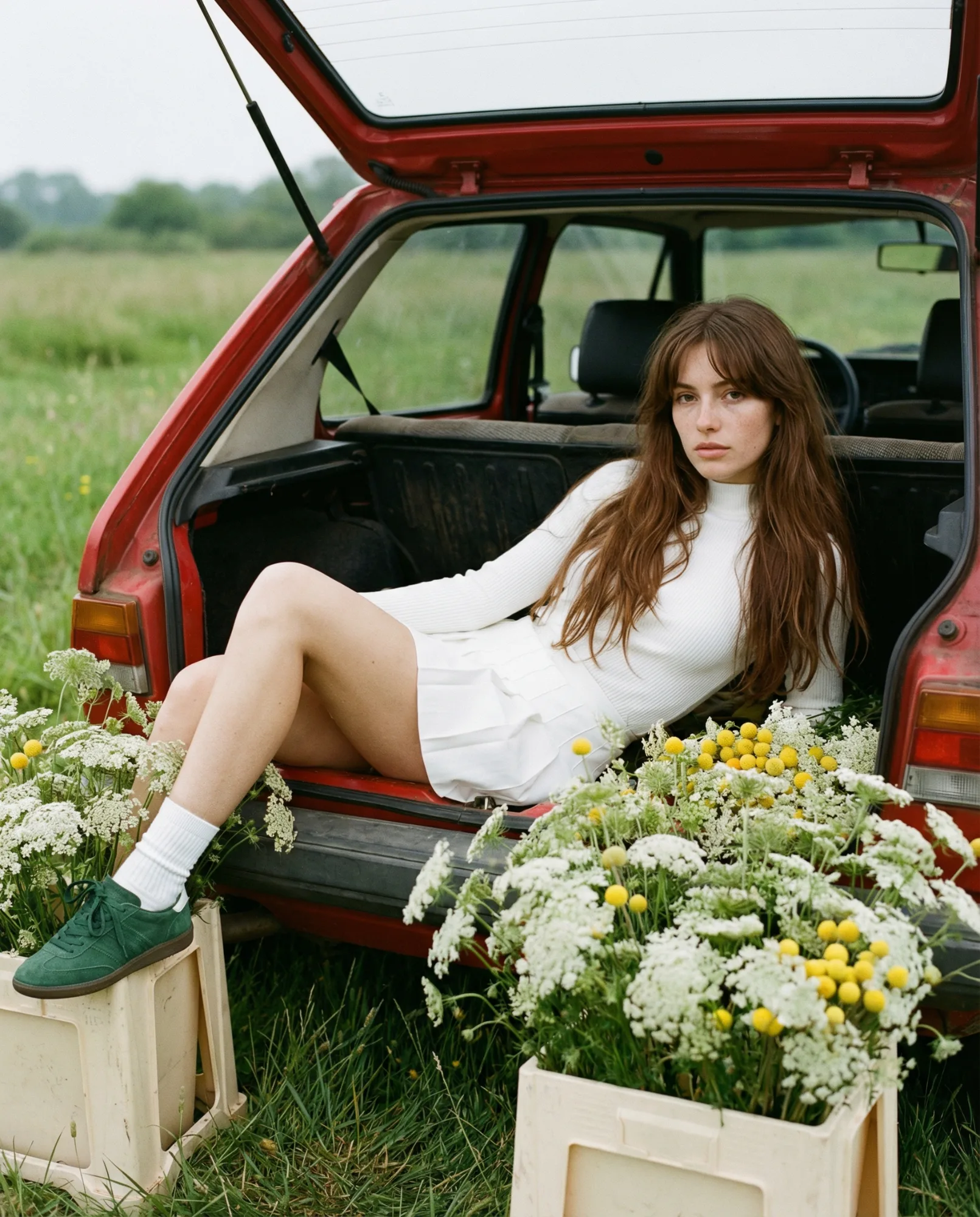 Donna seduta nel bagagliaio di un’auto rossa circondata da cassette di fiori in un campo rurale.