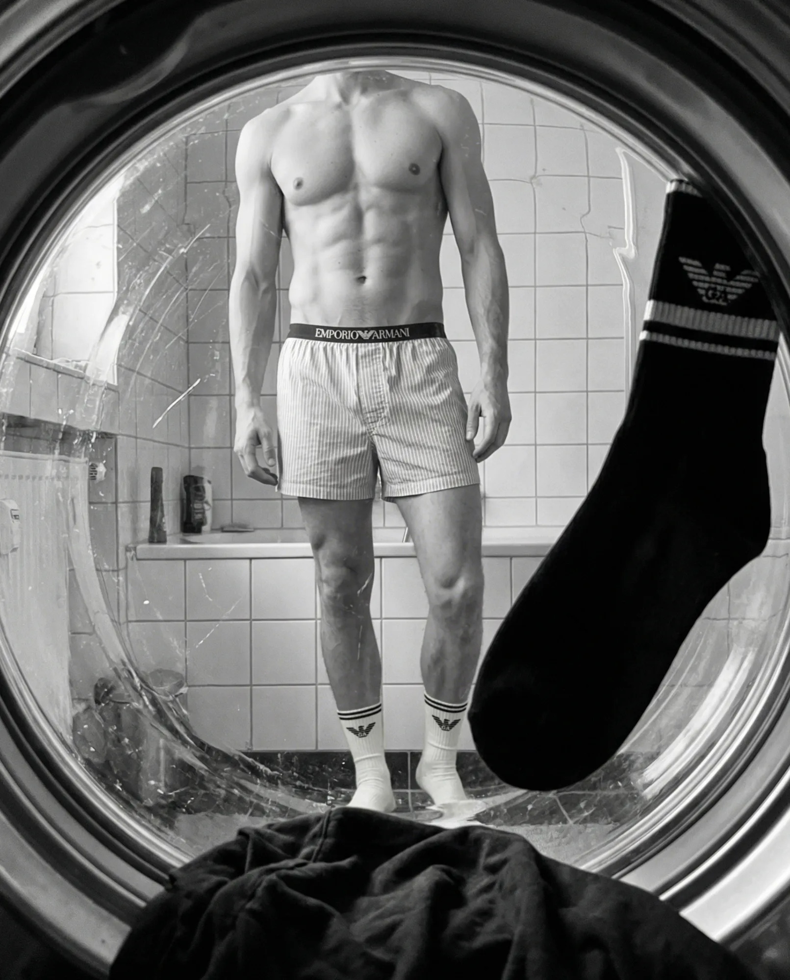 Vista dall’interno di una lavatrice: uomo in piedi in bagno con boxer e calzini Emporio Armani mentre un calzino scuro cade verso il cestello, fotografia fashion in bianco e nero.