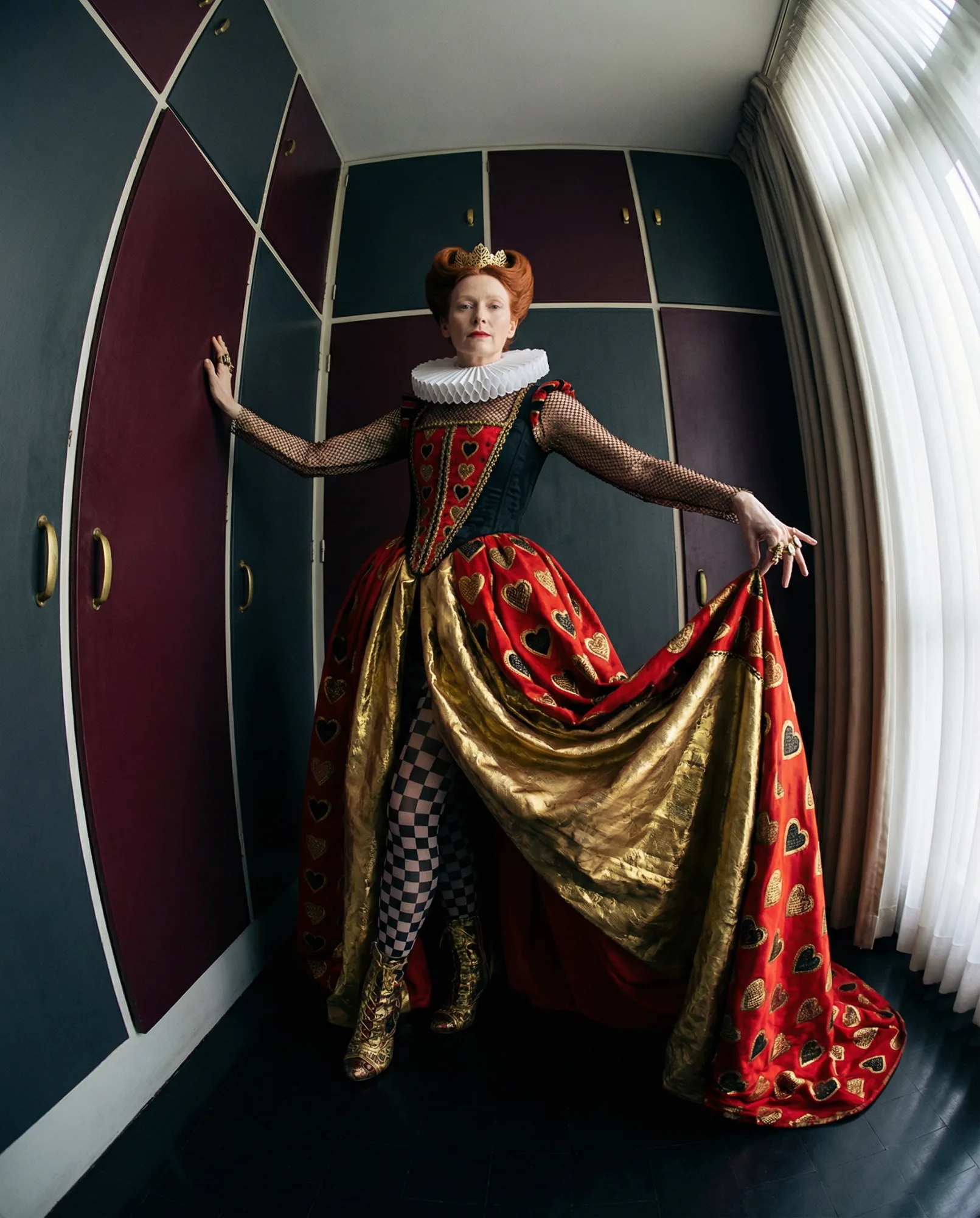 Donna in abito teatrale ispirato alla Regina di Cuori con corsetto nero, grande colletto bianco e ampia gonna rossa decorata con cuori dorati, posa in una stanza geometrica con pareti colorate e luce naturale dalla finestra.