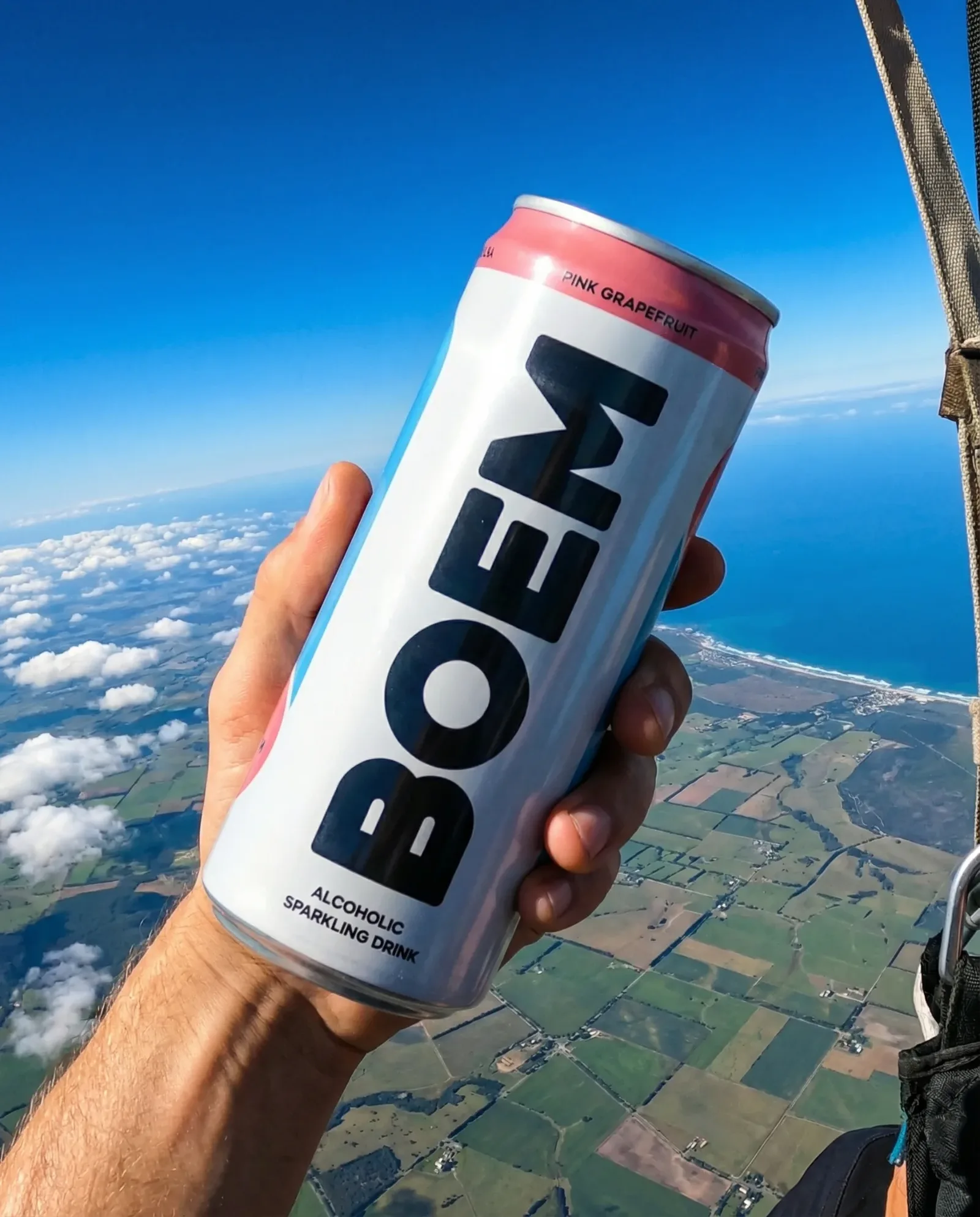 Lattina di bevanda frizzante tenuta in mano con vista aerea su campi e costa sotto un cielo limpido.