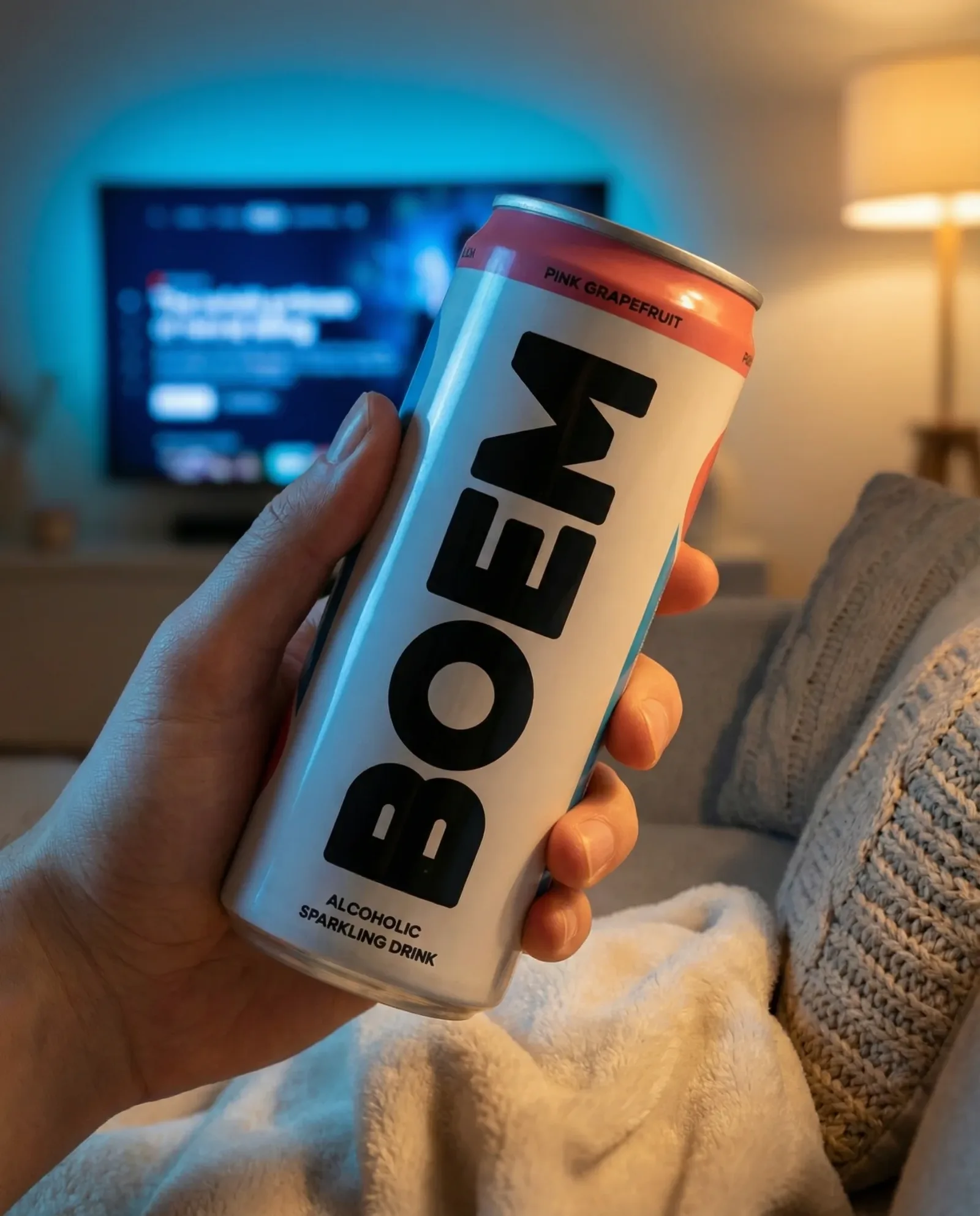 Lattina di bevanda frizzante tenuta in mano in un salotto serale, con televisione accesa e atmosfera rilassata.