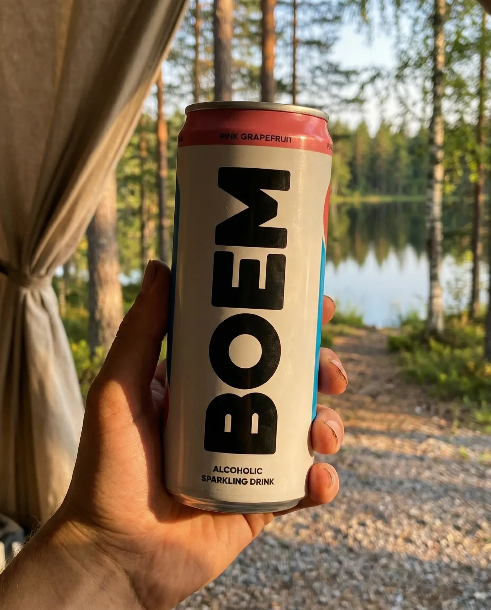 Mano che tiene una lattina di bevanda frizzante davanti a un lago immerso nel bosco, illuminato dalla luce calda del tramonto.