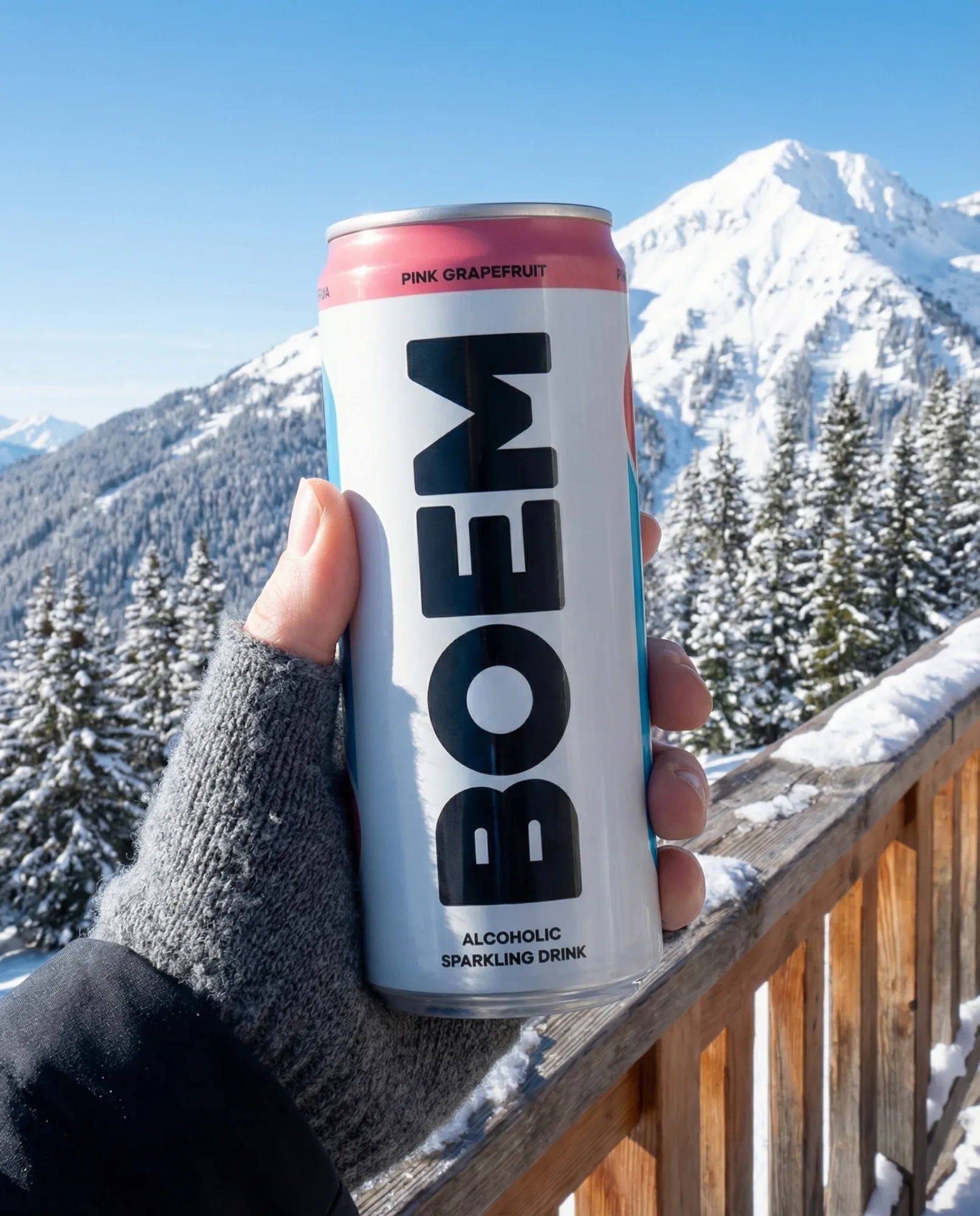 Mano con guanto invernale che tiene una lattina di bevanda frizzante davanti a un panorama alpino con montagne innevate e cielo limpido.