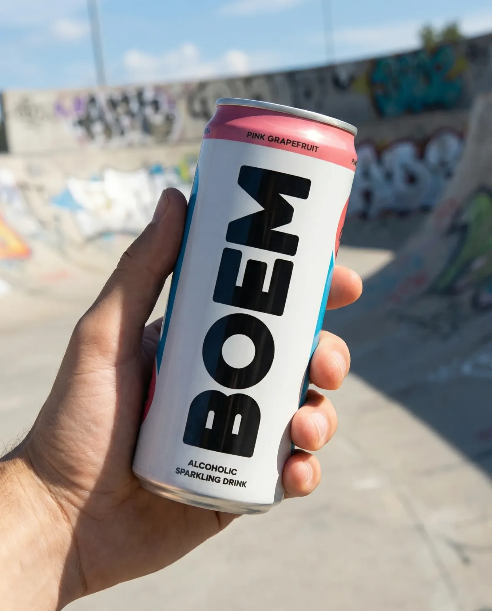 Lattina di bevanda frizzante analcolica tenuta in mano in uno skatepark all’aperto con rampe e graffiti sullo sfondo.