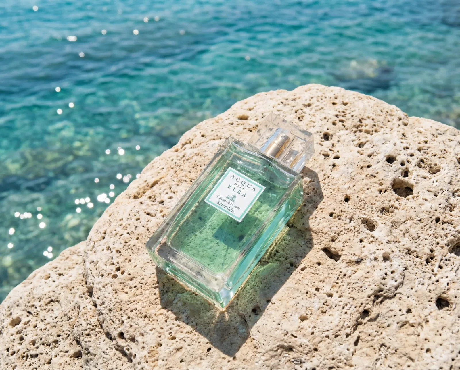 Bottiglia di profumo verde acqua su roccia chiara con mare cristallino sullo sfondo.