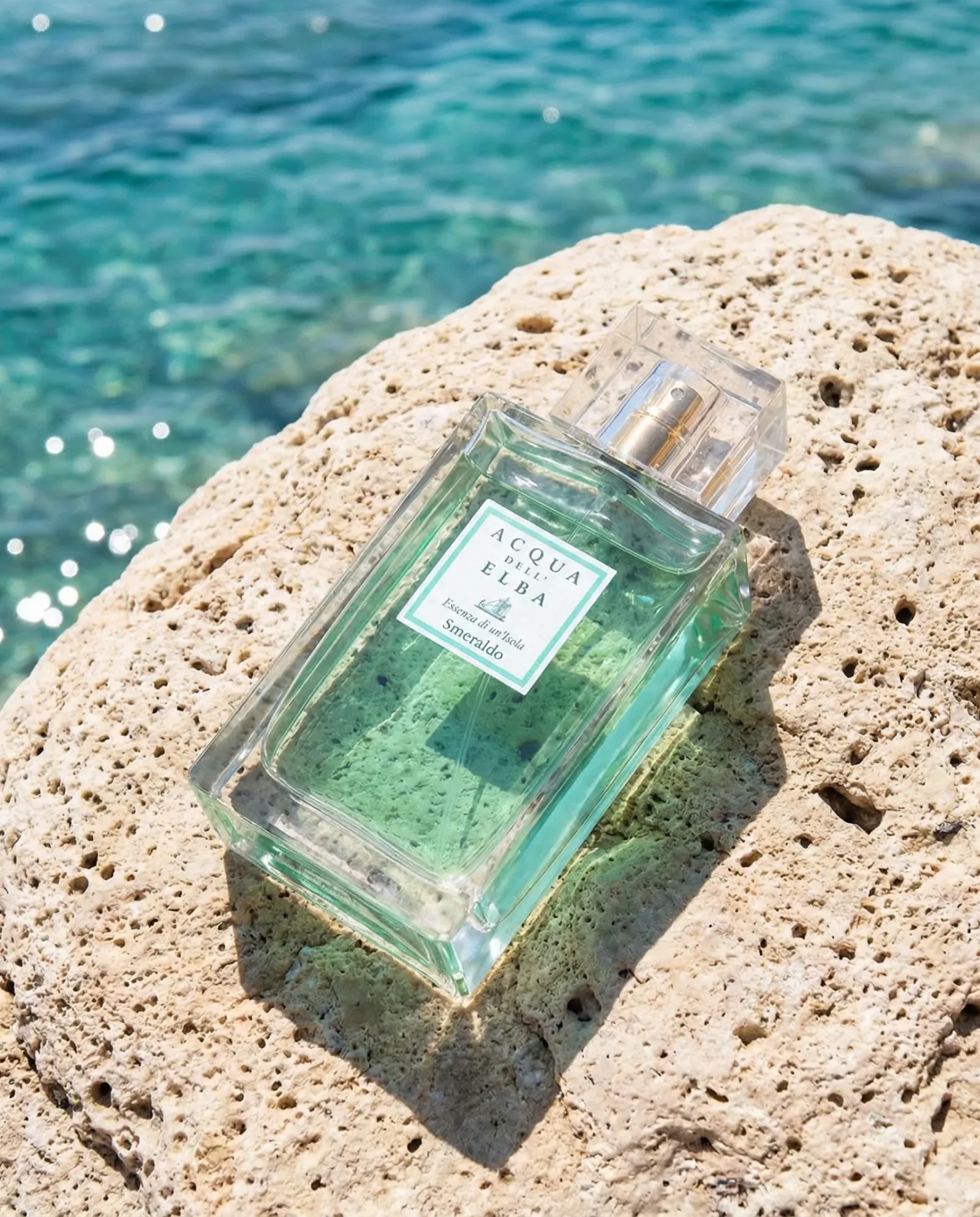 Bottiglia di profumo verde acqua su roccia chiara con mare cristallino sullo sfondo.