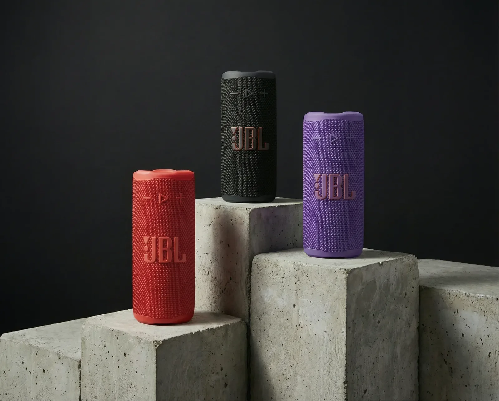 Tre speaker portatili colorati su piedistalli in cemento con sfondo scuro.
