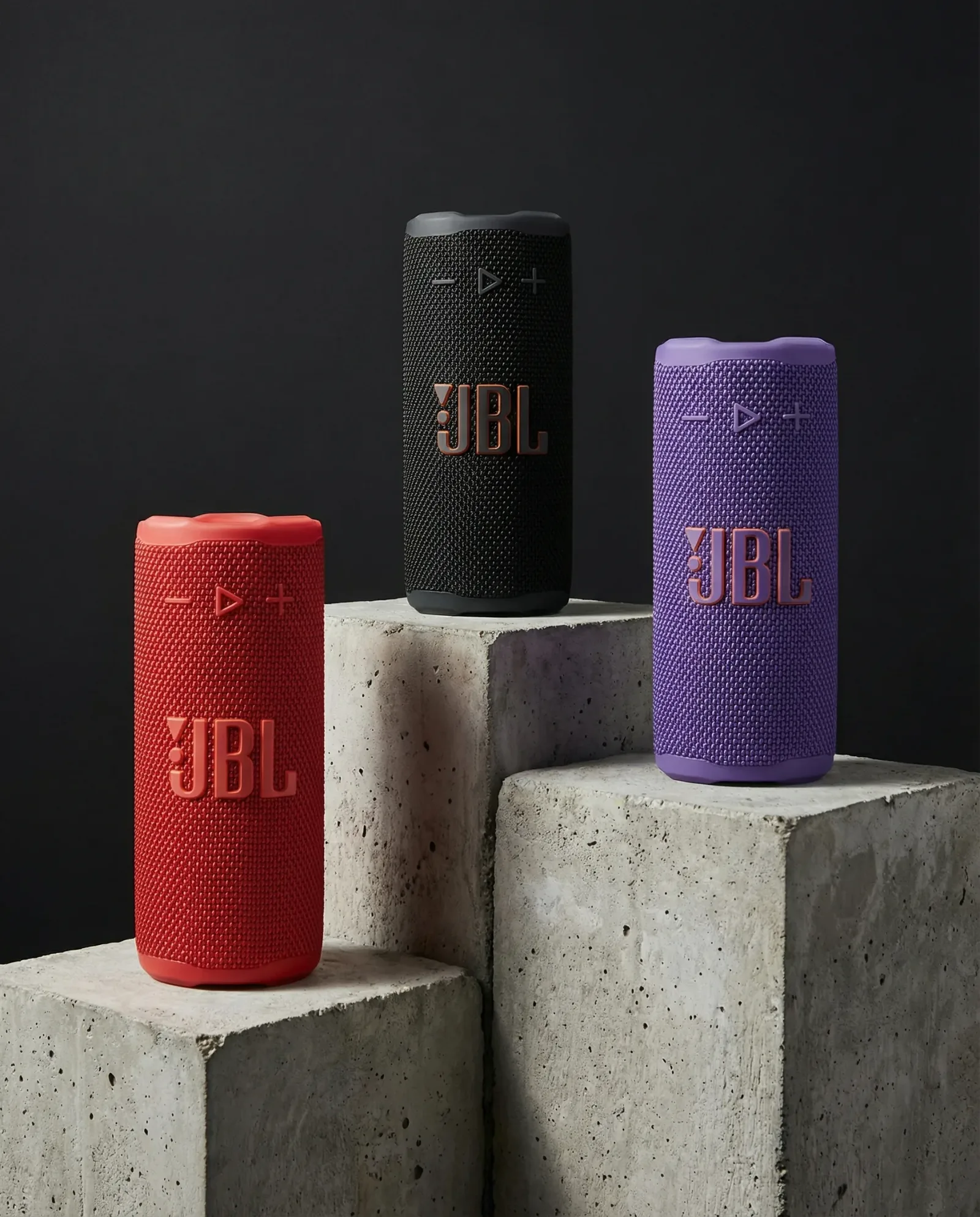 Tre speaker portatili colorati su piedistalli in cemento con sfondo scuro.