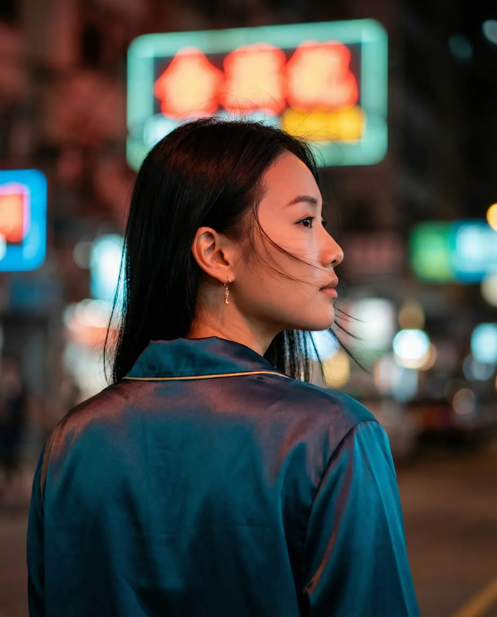 Ritratto di profilo di una donna asiatica con capelli lunghi neri e camicia satinata blu petrolio in una strada urbana notturna con luci al neon e bokeh colorati sullo sfondo.