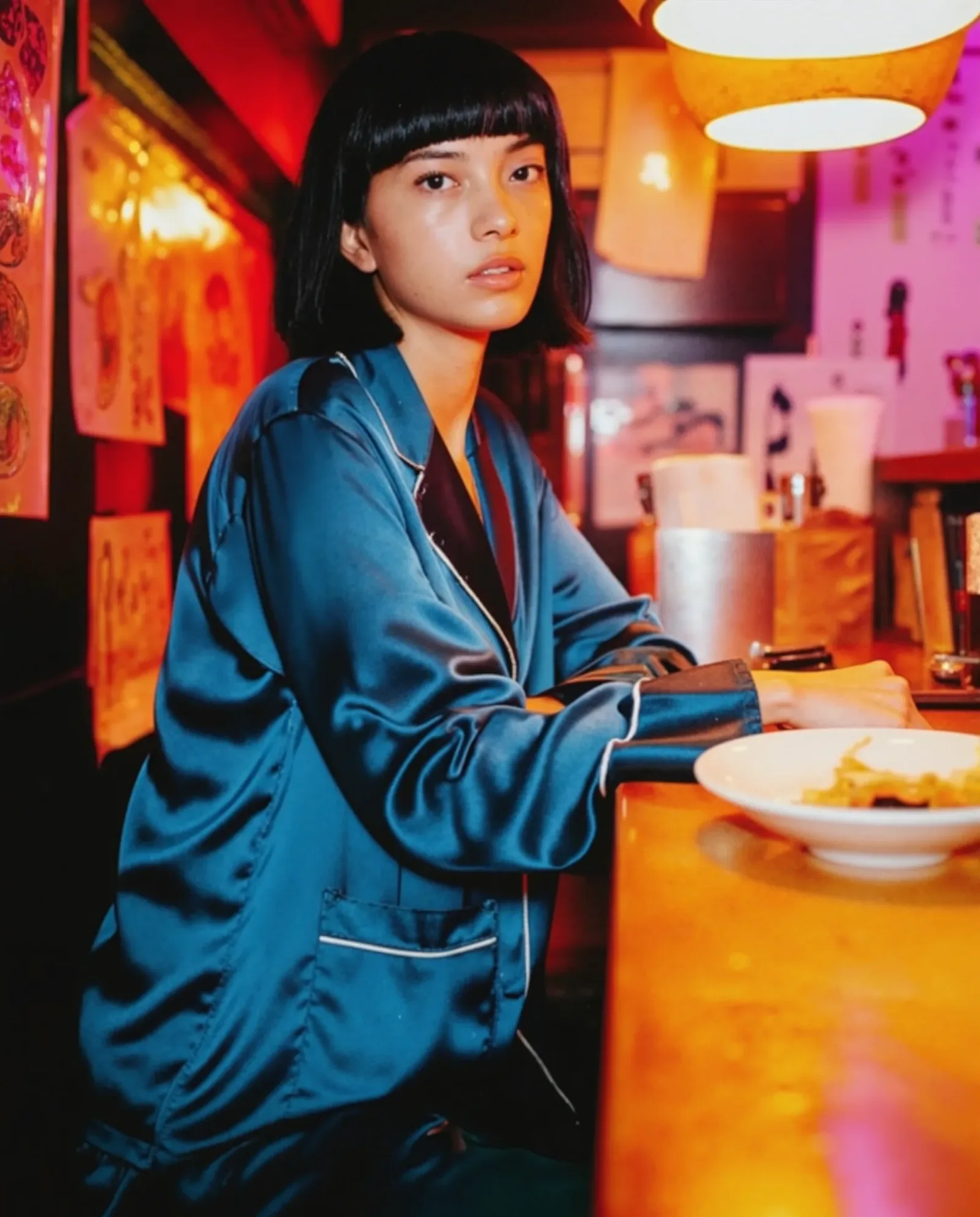 Modella asiatica con capelli corti neri e frangia seduta al bancone di un ristorante asiatico illuminato da luci rosse e arancioni, indossa una camicia satinata blu petrolio in un’atmosfera cinematografica notturna.