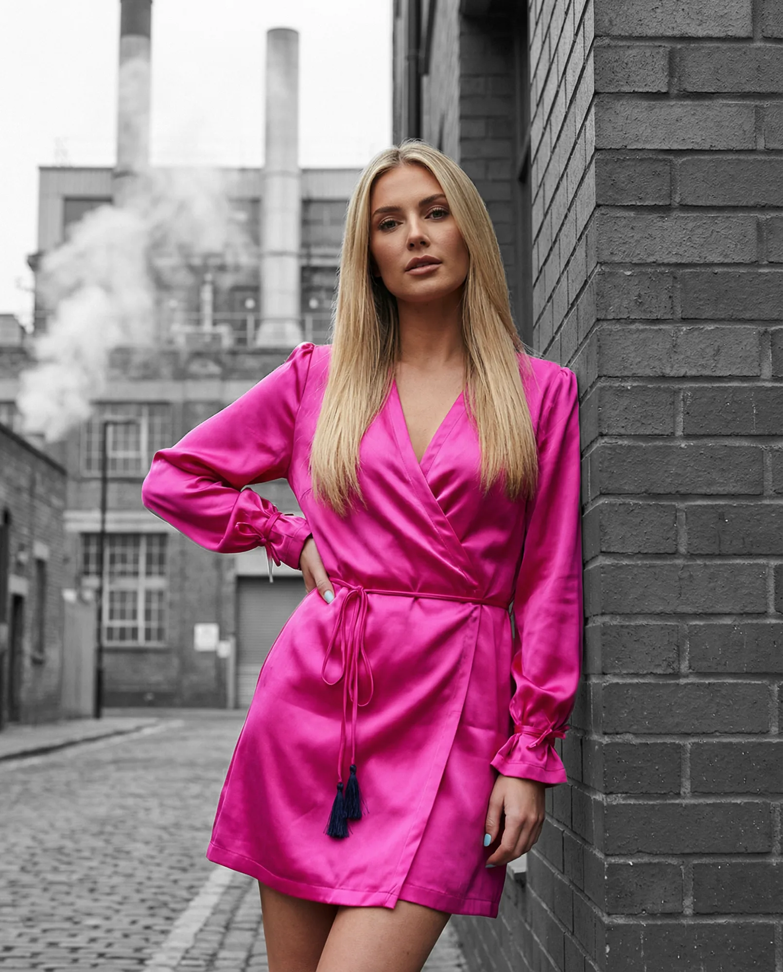 Modella bionda con abito satinato fucsia in una strada industriale urbana, con edifici e ciminiere sullo sfondo in bianco e nero.