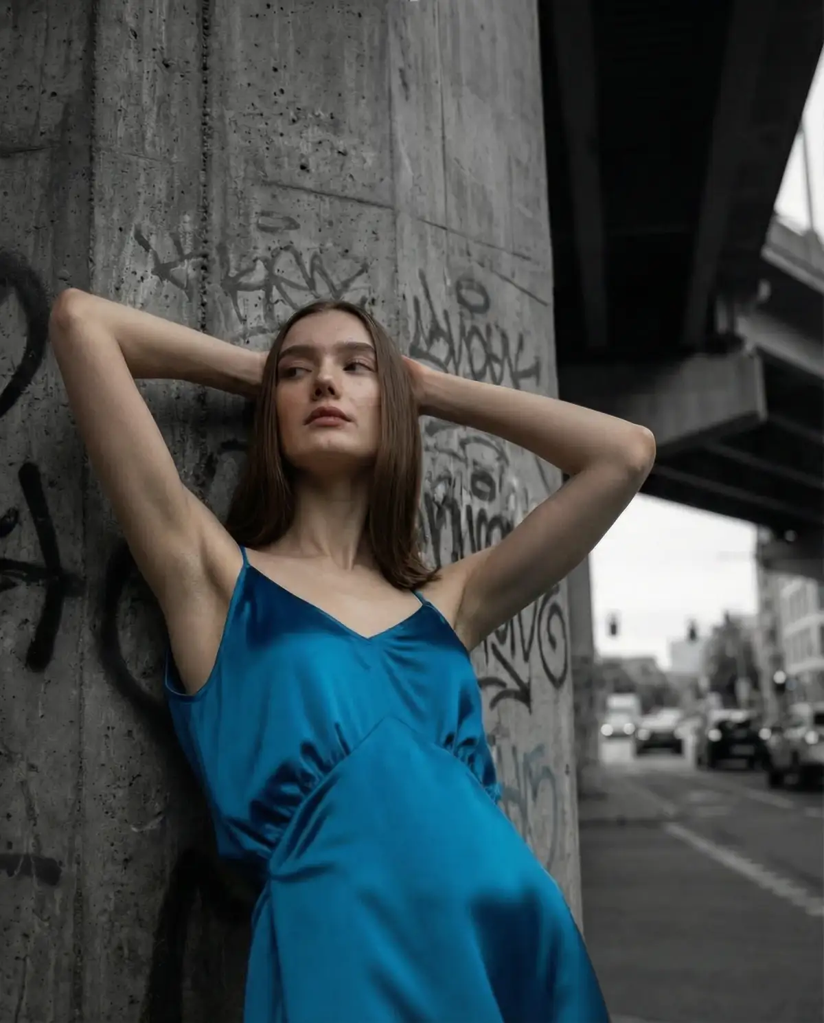 Modella con abito satinato blu petrolio appoggiata a un pilastro di cemento con graffiti sotto un ponte urbano, ritratto fashion con sfondo urbano in bianco e nero.